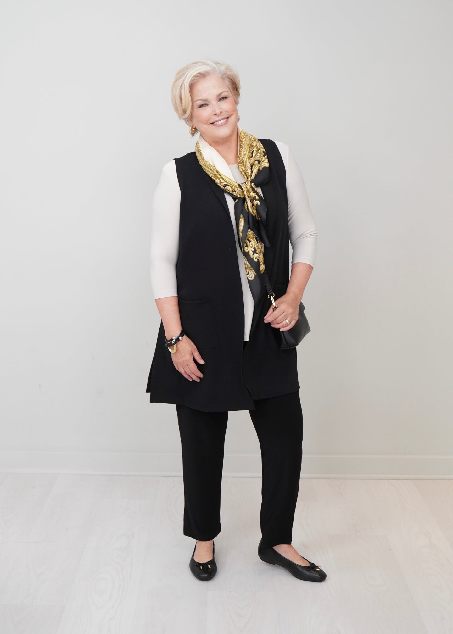 An woman stands in a studio wearing a black sleeveless vest over a white long-sleeve top, black pants, black flats, and a black-and-gold patterned scarf. She holds a small black clutch against a light gray wall and pale floor.
