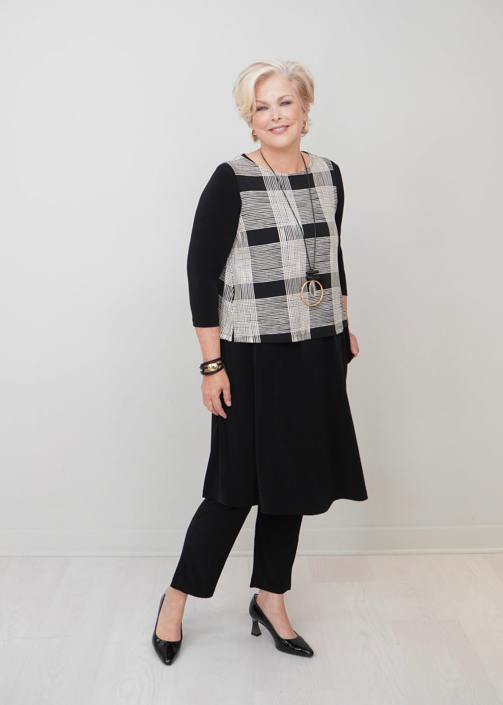An older woman modeling a black, white, and gray plaid vest over a black three-quarter sleeve top, black pants, and black heels, standing against a light gray studio background.