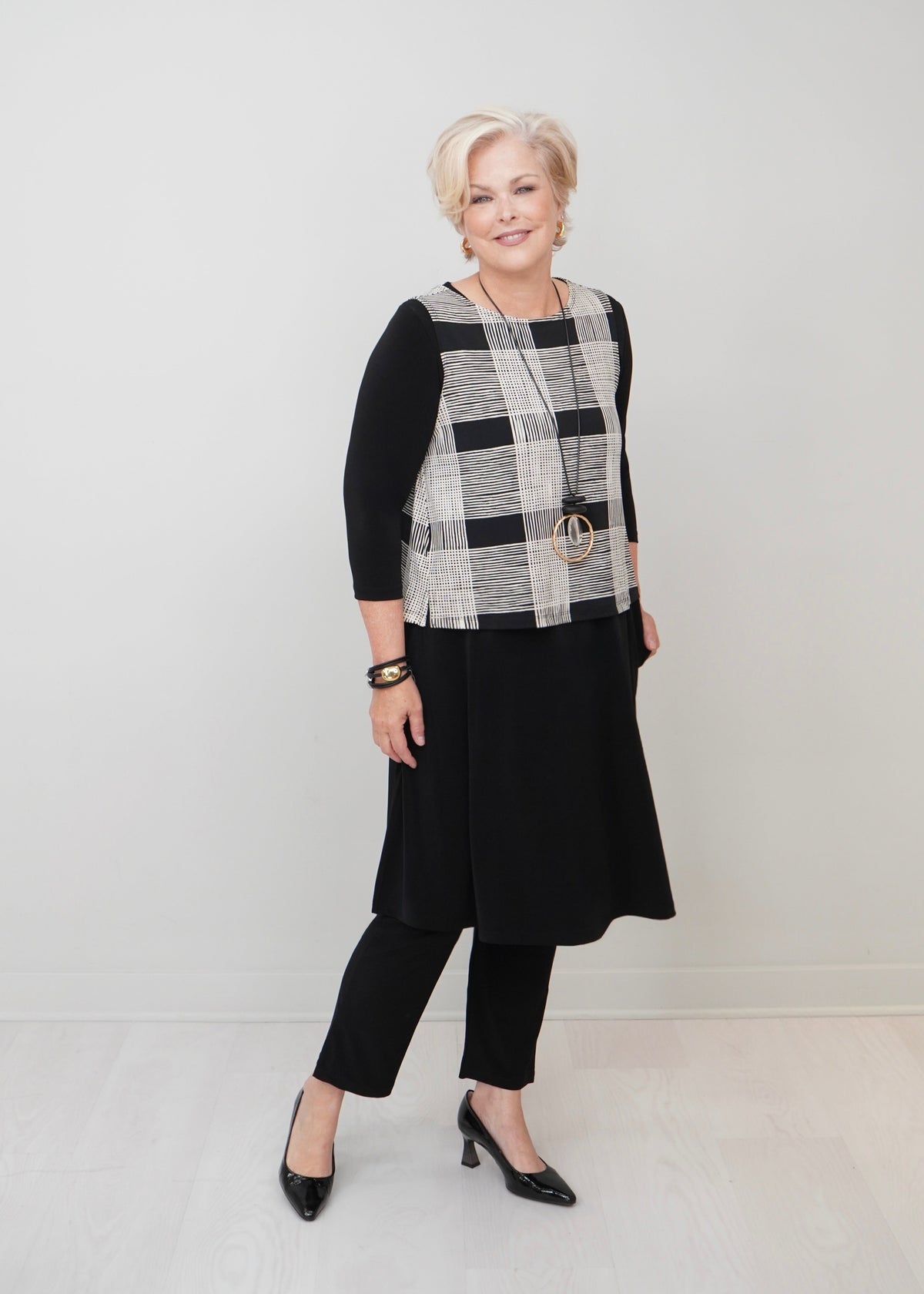 An older woman modeling a black, white, and gray plaid vest over a black three-quarter sleeve top, black pants, and black heels, standing against a light gray studio background.