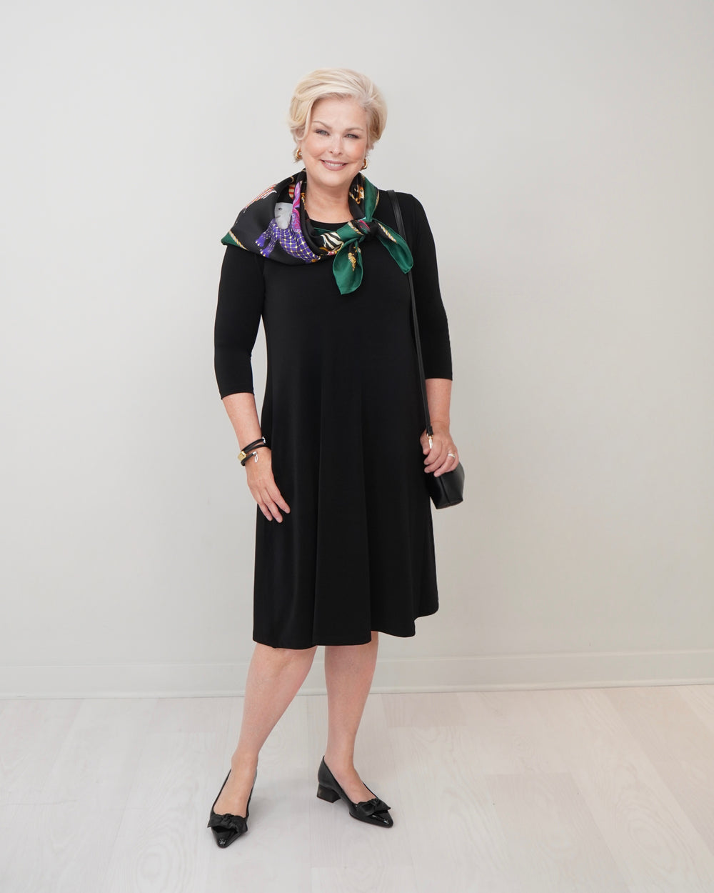 A woman modeling a black knee-length dress with three-quarter sleeves, styled with a green and purple floral patterned scarf, black pointed-toe heels, and a small black clutch, set against a plain light gray studio background.
