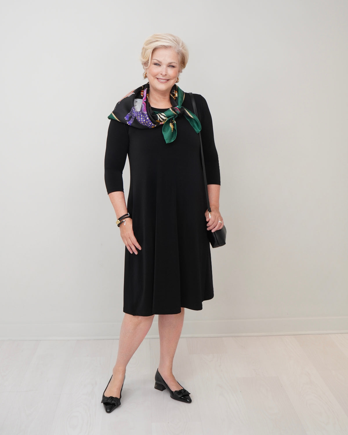 A woman modeling a black knee-length dress with three-quarter sleeves, styled with a green and purple floral patterned scarf, black pointed-toe heels, and a small black clutch, set against a plain light gray studio background.