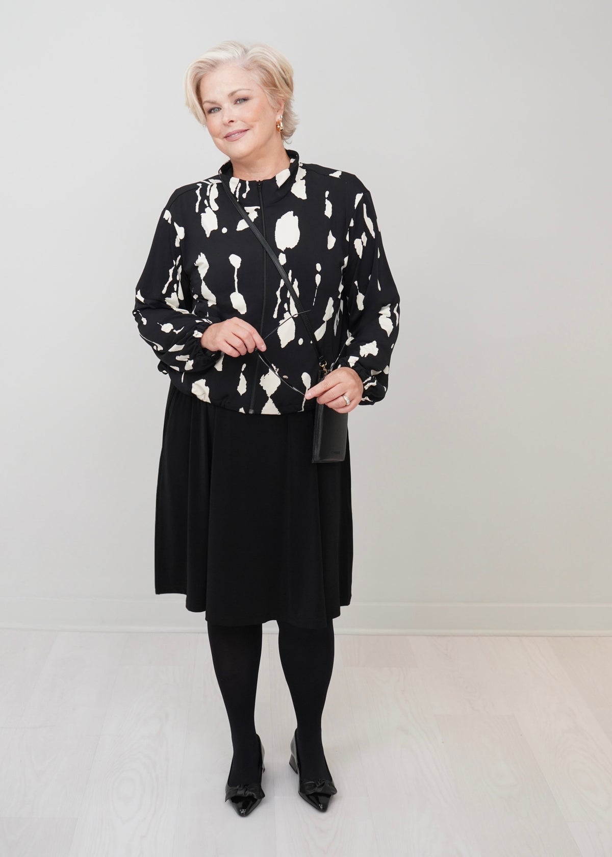 An woman stands in a minimalist studio wearing a black and white abstract patterned long-sleeve top, a black midi skirt, black tights, and black pointed shoes, holding a small black handbag.