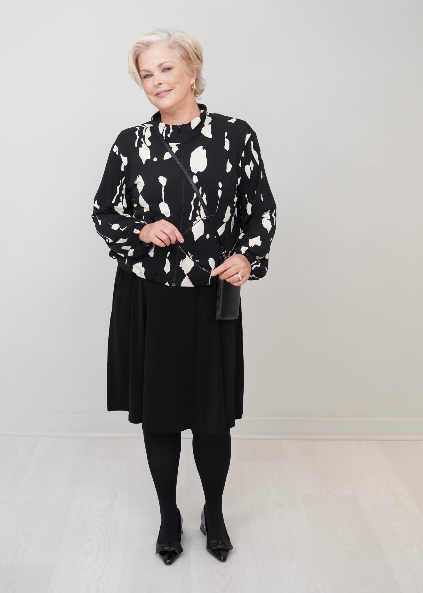 An woman stands in a minimalist studio wearing a black and white abstract patterned long-sleeve top, a black midi skirt, black tights, and black pointed shoes, holding a small black handbag.