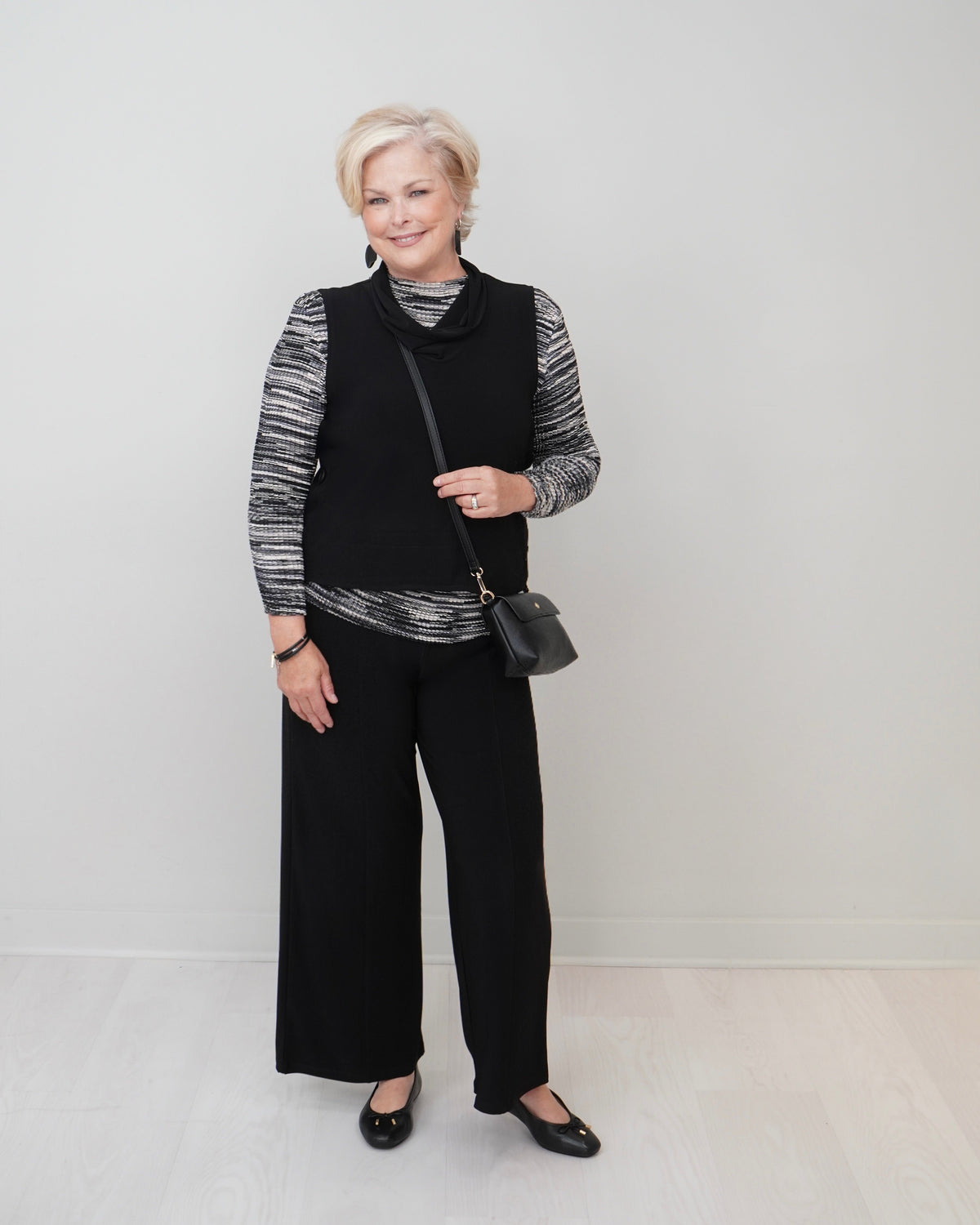 A woman stands in a studio wearing a black sleeveless vest over a black-and-white striped long-sleeve top, black wide-leg pants, and black flats. She is holding a small black shoulder bag against a plain light gray wall and pale floor.