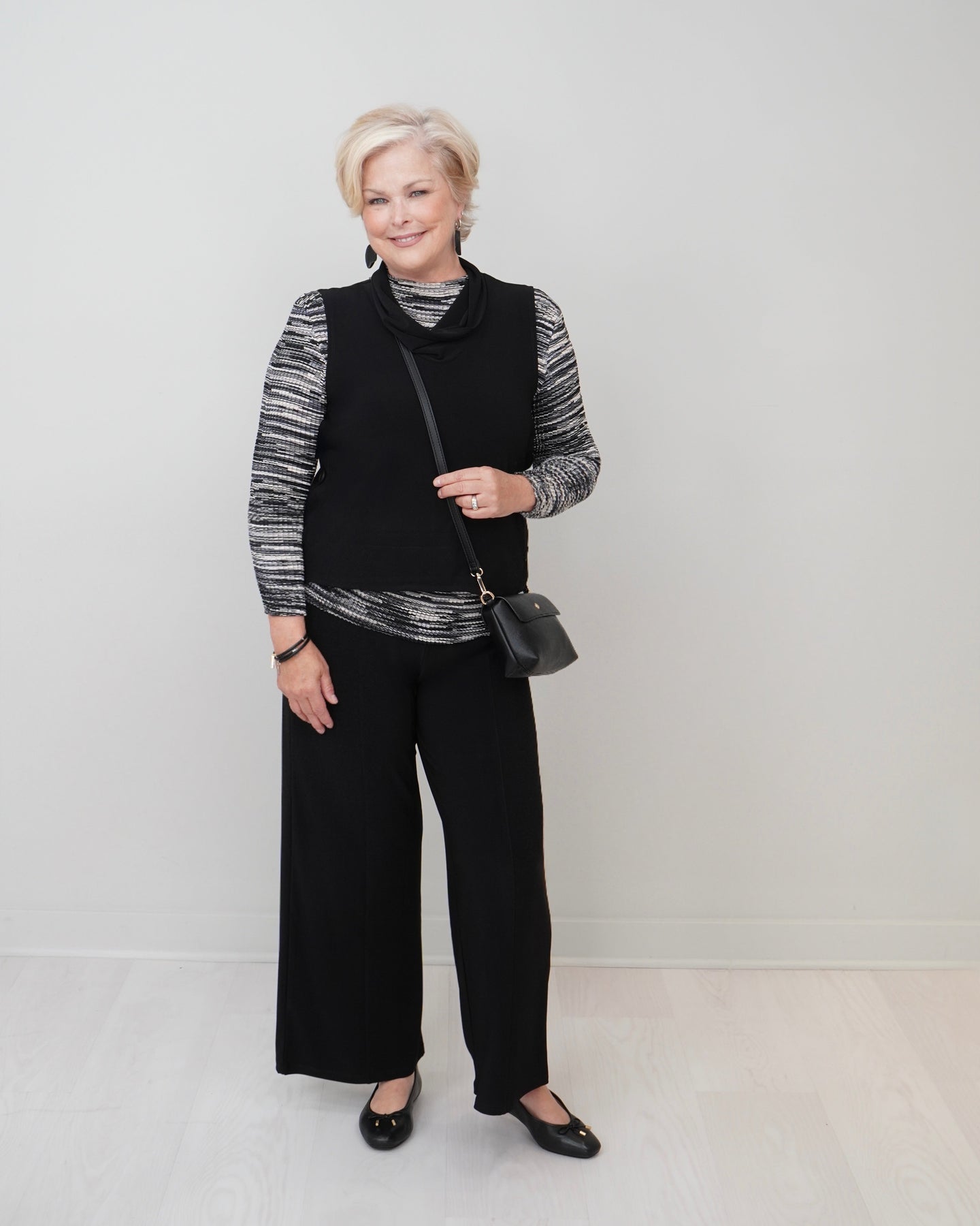 A woman stands in a studio wearing a black sleeveless vest over a black-and-white striped long-sleeve top, black wide-leg pants, and black flats. She is holding a small black shoulder bag against a plain light gray wall and pale floor.
