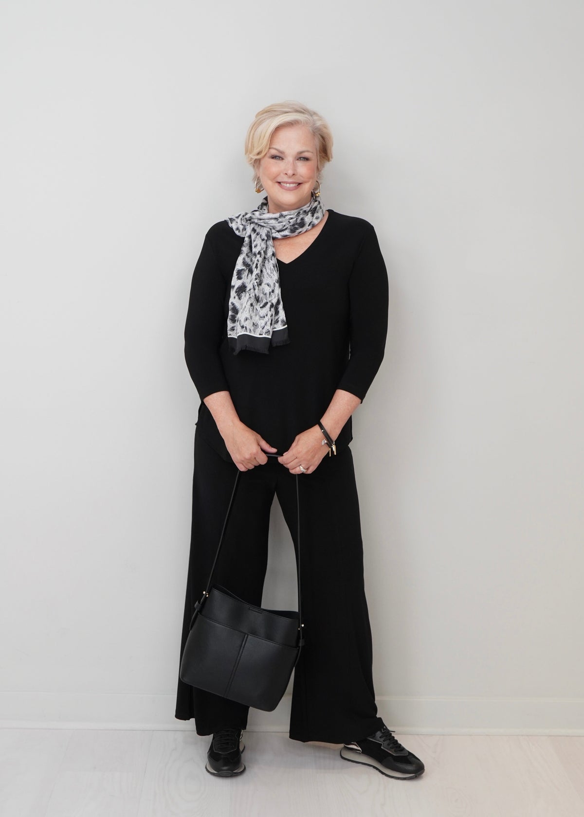 A woman stands against a plain light gray wall wearing a black long-sleeve top and wide-leg pants, with a black-and-white patterned scarf and a black shoulder bag.