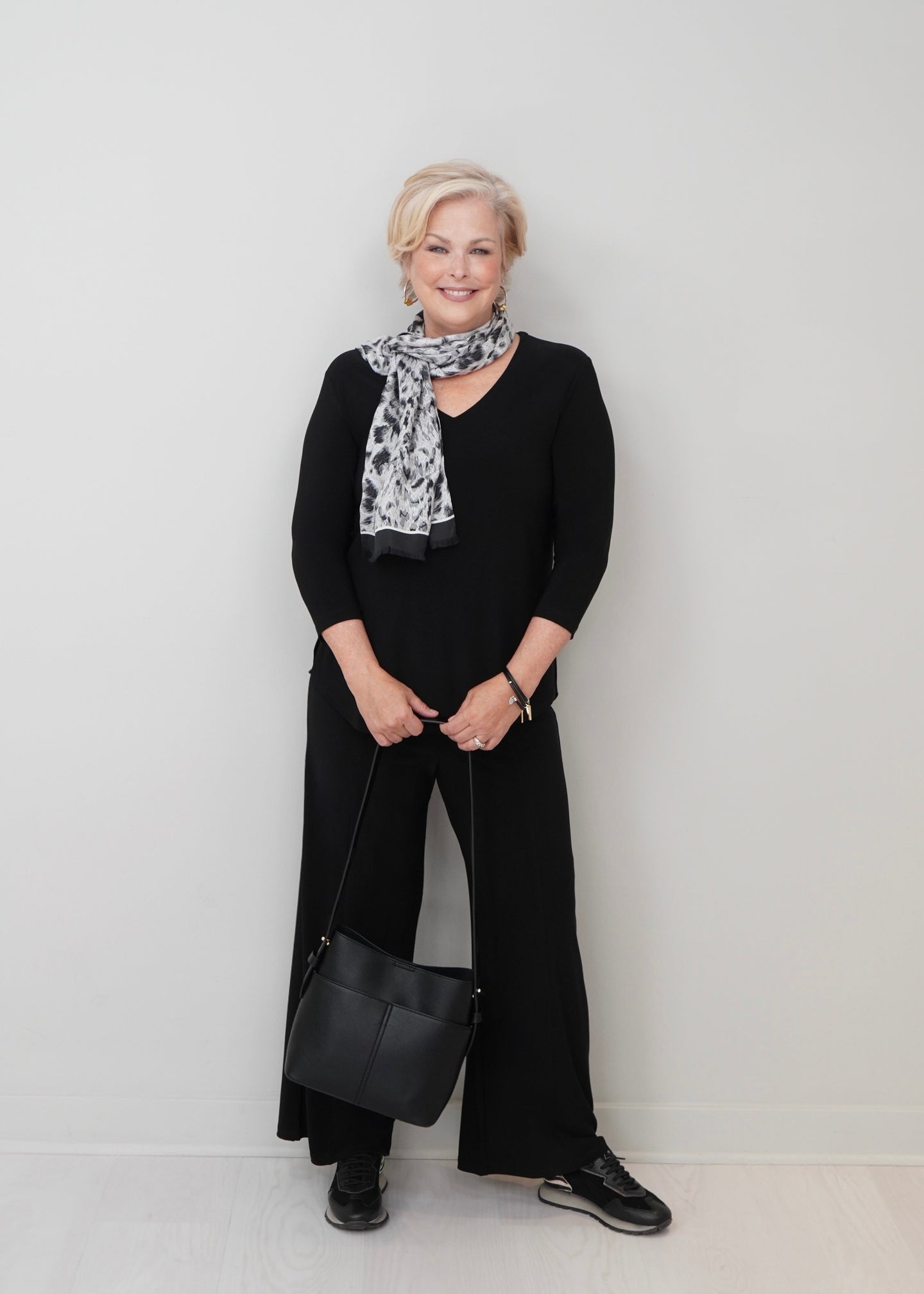 A woman stands against a plain light gray wall wearing a black long-sleeve top and wide-leg pants, with a black-and-white patterned scarf and a black shoulder bag.