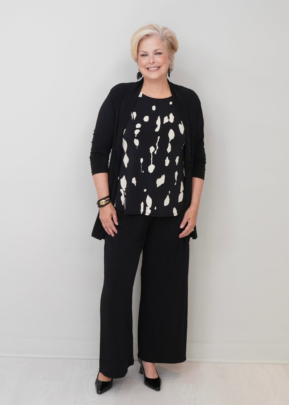 A woman modeling a black cardigan, black wide-leg pants, and a black top with a white abstract pattern, standing against a plain light gray wall.