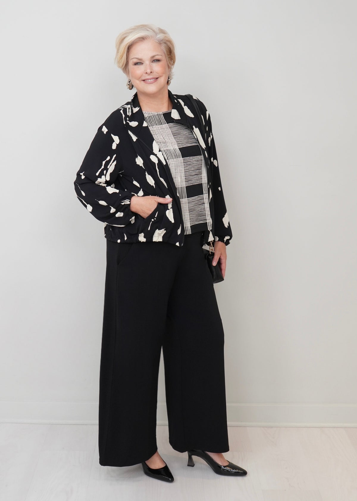 An older woman stands in a studio wearing a black and white abstract print jacket over a black and white plaid top, paired with black wide-leg pants and black pointed heels.