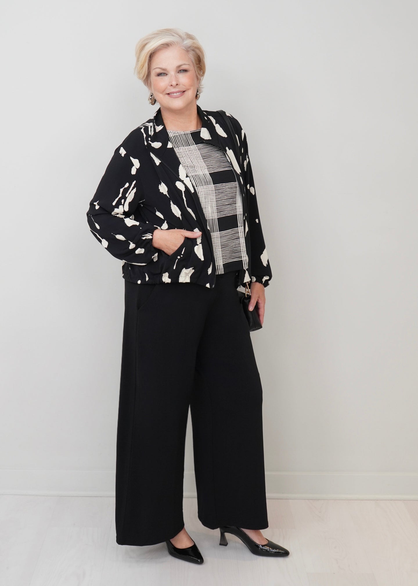 An older woman stands in a studio wearing a black and white abstract print jacket over a black and white plaid top, paired with black wide-leg pants and black pointed heels.