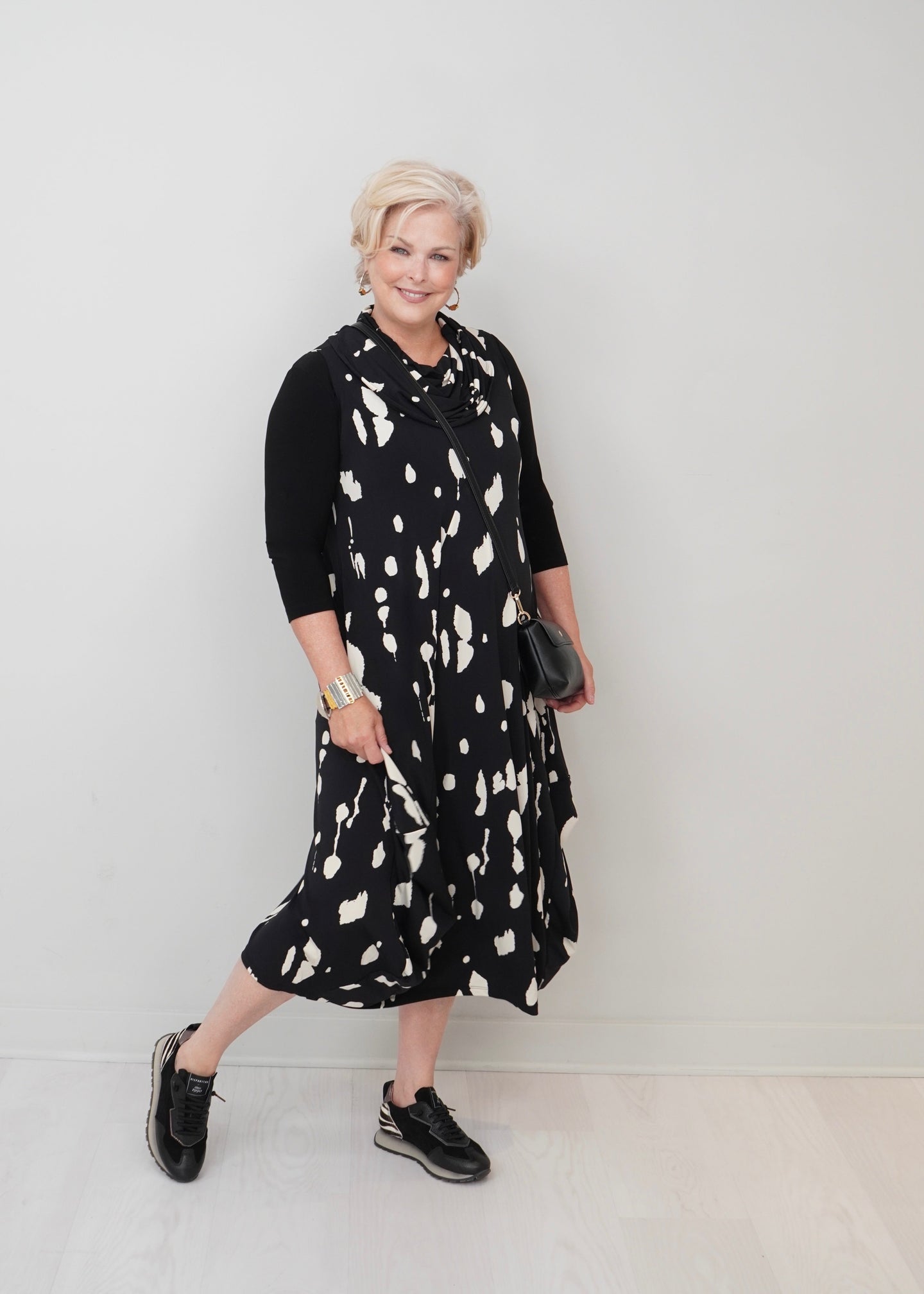 An woman with short blonde hair models a black and white abstract patterned midi dress over a black three-quarter sleeve top, styled with black sneakers and a small black handbag against a plain light gray wall.